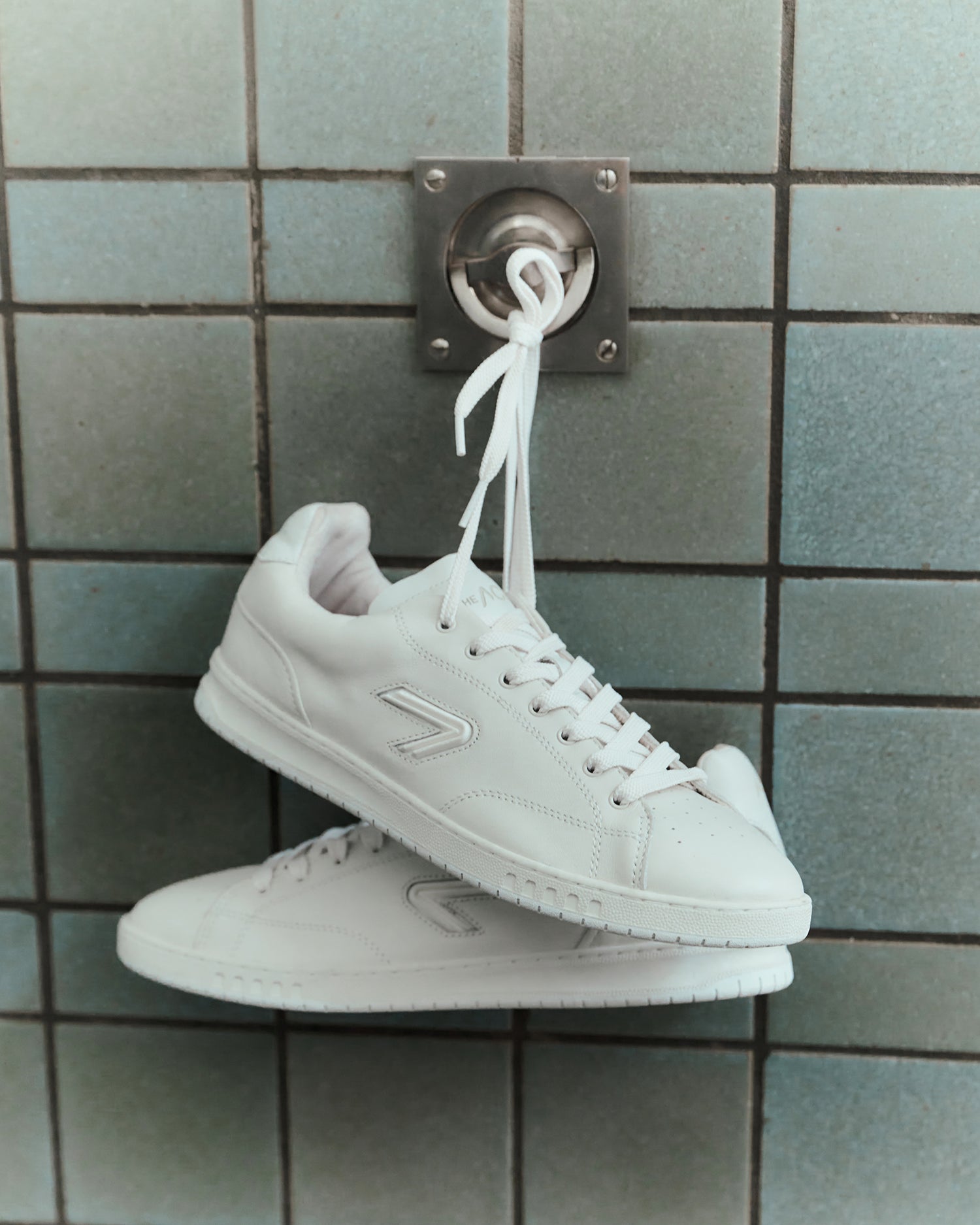 The ACE Hub Shot sneakers in off-white, hanging by laces against tiled wall

